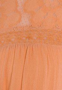 Pfirsichfarbener Spitzenstoff mit floraler Stickerei am Ausschnitt, kombiniert mit weichem, plissiertem Material im Rock, das eine gestaffelte Textur erzeugt.