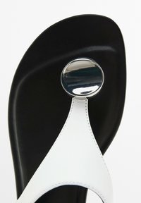 Hvide thong sandaler med en skinnende rund metallaccent på en sort sål, der har strukturerede hvide læderstropper og syet detaljering.