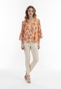Blomstret bluse i orange og creme med klokkeærmer, parret med beige tætsiddende bukser og nude sandaler med straps, mod en hvid baggrund.