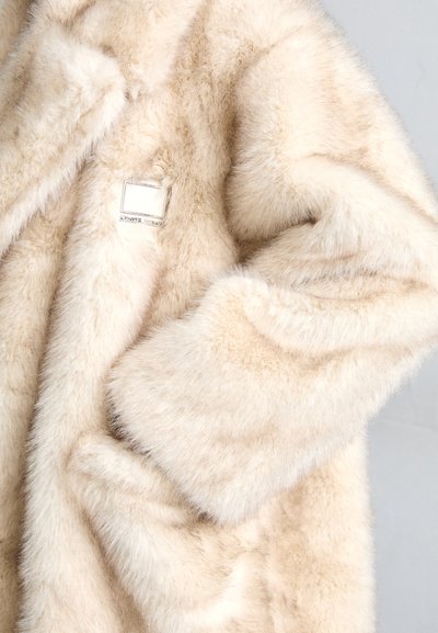 Dirbtinio kailio paltas švelniai kreminės spalvos, su puriu paviršiumi. Pasižymi kvadratine etikete ir laisvu siluetu, demonstruojančiu besiūlį dizainą.
