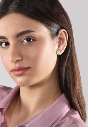 Grüne Ohrstecker mit strukturiertem, hellgrünem Mittelpunkt, umgeben von einem silberfarbenen Rand. Modell trägt ein rosa Hemd mit Kragen.