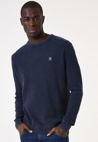 Mann trägt einen navyblauen Waffelpullover mit kleinem Brustlogo, die Hände in den Taschen, steht vor einem schlichten hellen Hintergrund.