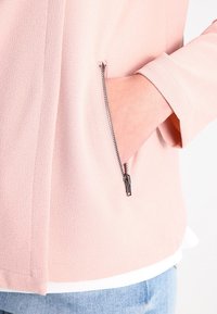 Hand in open zipper pocket of light pink textured jacket worn over white shirt and blue jeans against a white background.