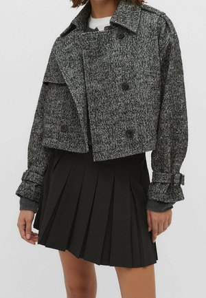 Vrouw die een grijze, cropt herringbone mantel met dubbele rij knopen en een zwarte geplooide rok draagt, staand tegen een effen achtergrond.