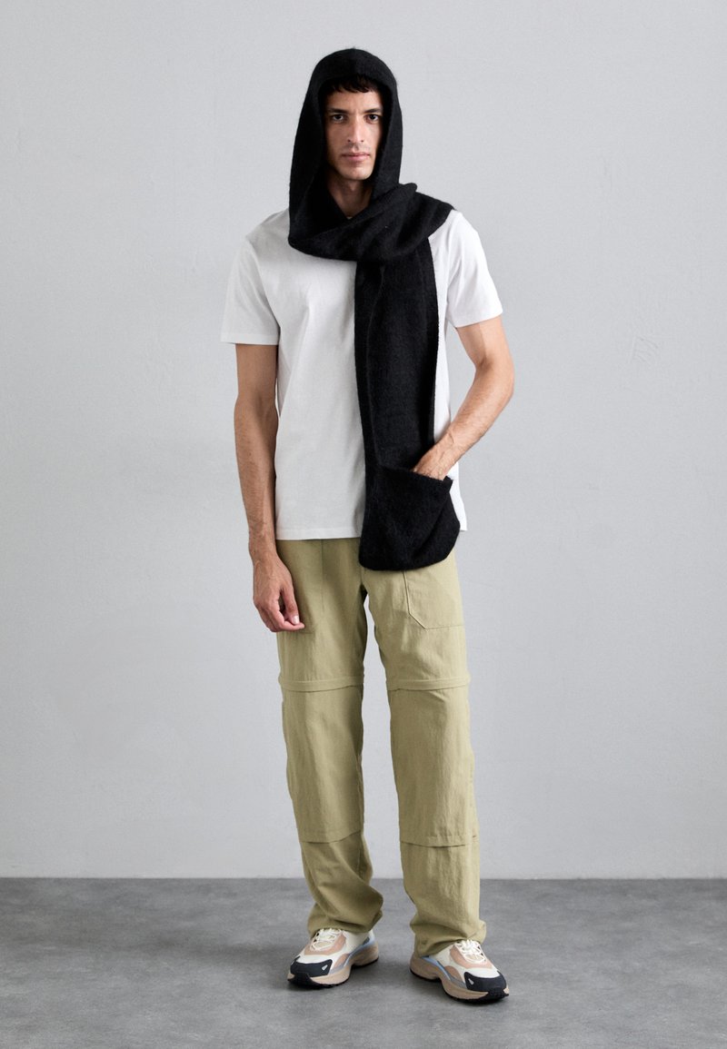 Écharpe noire portée comme une capuche, avec une texture douce ; associée à un t-shirt blanc et un pantalon cargo vert olive, complétée par des chaussures de sport.