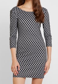 Czarna i biała sukienka w geometryczny wzór, z rękawami do trzech czwartych i szerokim dekoltem, wykonana z elastycznego materiału.
