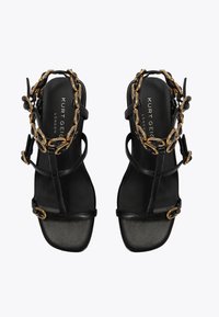 Czarne sandały na obcasie z wieloma paskami, złotym łańcuchem jako akcentem oraz gładką podeszwą. Posiadają markowy wzór wkładki i minimalistyczny kształt.