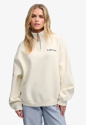 Femme portant un sweat-shirt crème à fermeture éclair mi-hauteur avec le logo S.DRY & Co. et un pantalon rayé bleu clair sur un fond blanc uni.