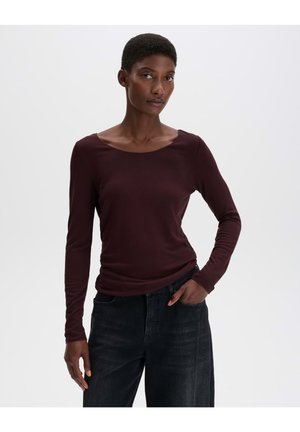 Femme aux cheveux courts portant une chemise bordeaux foncé à manches longues et un jean large foncé, debout avec une main dans la poche.