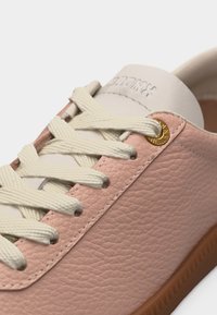 Κοντινό πλάνο ροζ δερμάτινου αθλητικού παπουτσιού με ανάγλυφη υφή, κρεμ κορδόνια, κρεμ γλώσσα, χρυσές τρύπες για κορδόνια και καφέ ελαστική σόλα.