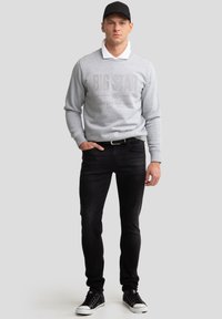 Mees, kes kannab musta nokamütsi, helerohelist dressipluusi, millel on tekst "BIG STAR", valget kraega särki, musti teksaseid ja musti tossusid, seisab halli tausta ees.