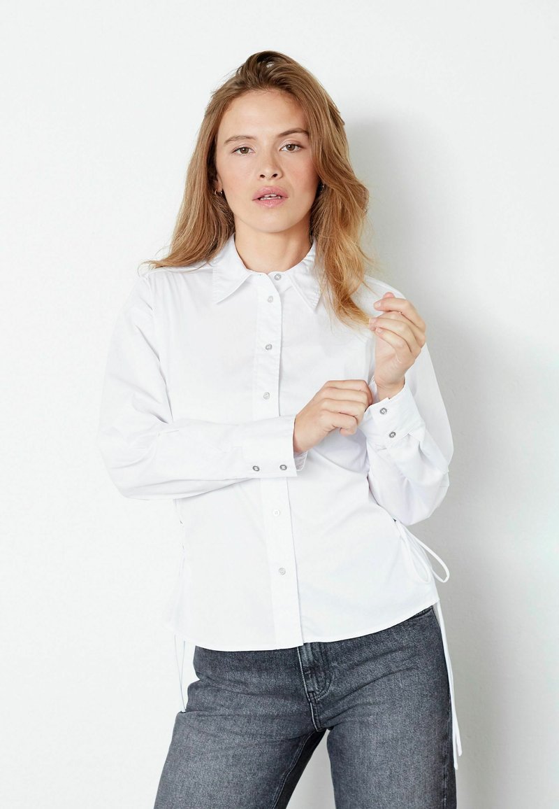 White button-up shirt with a collared neckline, long sleeves, and adjustable side ties. Made of smooth fabric with a fitted silhouette.