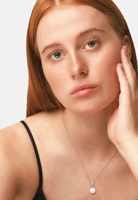 Collier en argent avec un pendentif en perle, porté par un mannequin aux longs cheveux roux, qui porte un haut noir, mettant en valeur une texture de peau lisse.