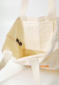 Sac fourre-tout en toile beige avec deux poignées, montrant l'intérieur vide et une petite poche intérieure avec une étiquette noire à l'intérieur.
