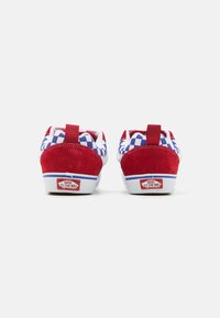 Sneaker in suede rosso e tessuto bianco con un motivo a scacchi blu sulla linguetta, dotate di un etichetta rossa sul tallone e logo visibile.