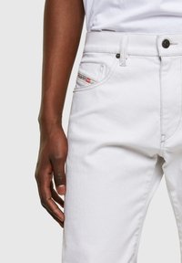 Dark-skinned hand resting beside white denim jeans with visible front pocket, belt loop, and branded fabric tag near the pocket.