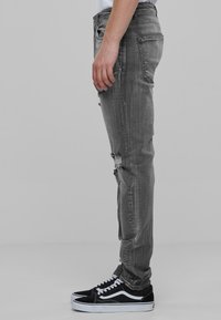 Jeans in denim grigio con vestibilità slim, caratterizzati da dettagli strappati e cuciture in vista. Abbinati a sneakers bianche e nere.