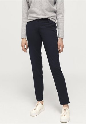 Persoon draagt slim-fit marineblauwe broek, lichtgrijze longsleeve en witte platformsneakers en staat voor een effen achtergrond.