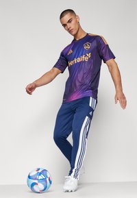Maglia sportiva viola con accenti gialli e maniche a righe, abbinata a pantaloni sportivi blu con righe bianche. Pallone da calcio accanto.