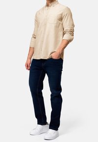 Chemise beige à manches longues avec boutons et une poche poitrine, associée à un jean bleu foncé et des baskets blanches. Design simple, texture lisse.