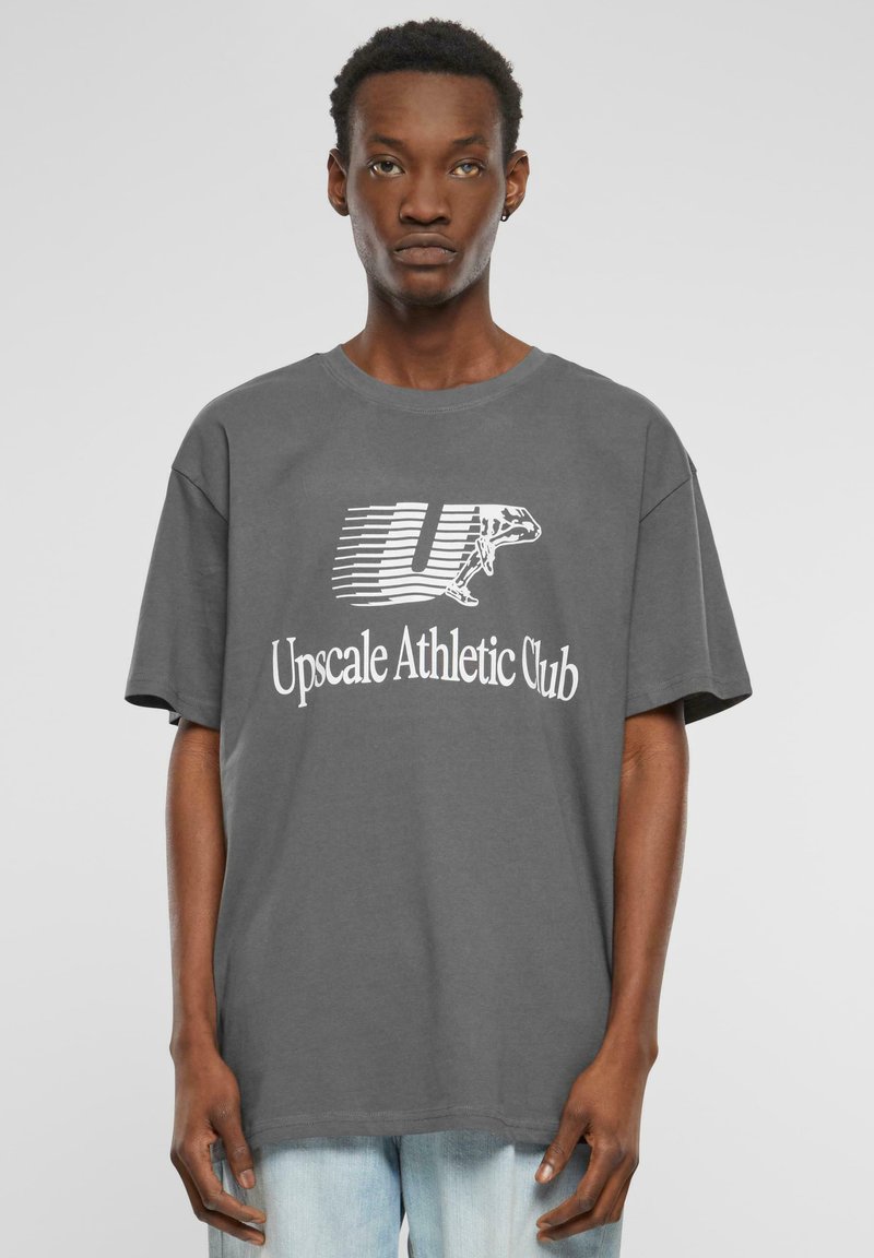 Šedé bavlněné tričko s krátkými rukávy, které obsahuje bílé grafické ztvárnění nápisů "Up" a "Upscale Athletic Club". Volný střih.