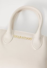 Bolso de mano de cuero crema con dos asas, que presenta un logo en relieve dorado y acentos cosidos en los bordes.