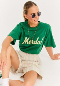 Zelené tričko s krátkym rukávom s krémovým nápisom "Morde!" v kombinácii s svetlo pruhovanou sukňou; okrúhle slnečné okuliare a farebný korálkový náhrdelník.