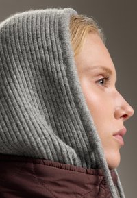 Szürke bordás kötött kapucni, amely részben eltakarja az arcot, egy bordó steppelt dzsekivel párosítva. A texturált anyagok réteges megjelenést kölcsönöznek.