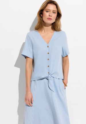 Kobieta ubrana w jasnoniebieski krótki top zapinany na guziki, wiązany w talii, z dopasowaną spódnicą z wysokim stanem, stoi na białym tle.