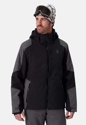 Homme portant une veste de ski noire et grise, un pantalon de ski gris, un bonnet blanc à motifs et des lunettes de ski sur le front, regardant légèrement sur le côté.