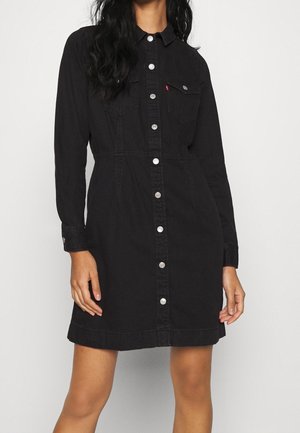 Mujer vistiendo un vestido de mezclilla negro, de manga larga y con botones al frente, con bolsillos delanteros y botones plateados, de pie frente a un fondo liso.