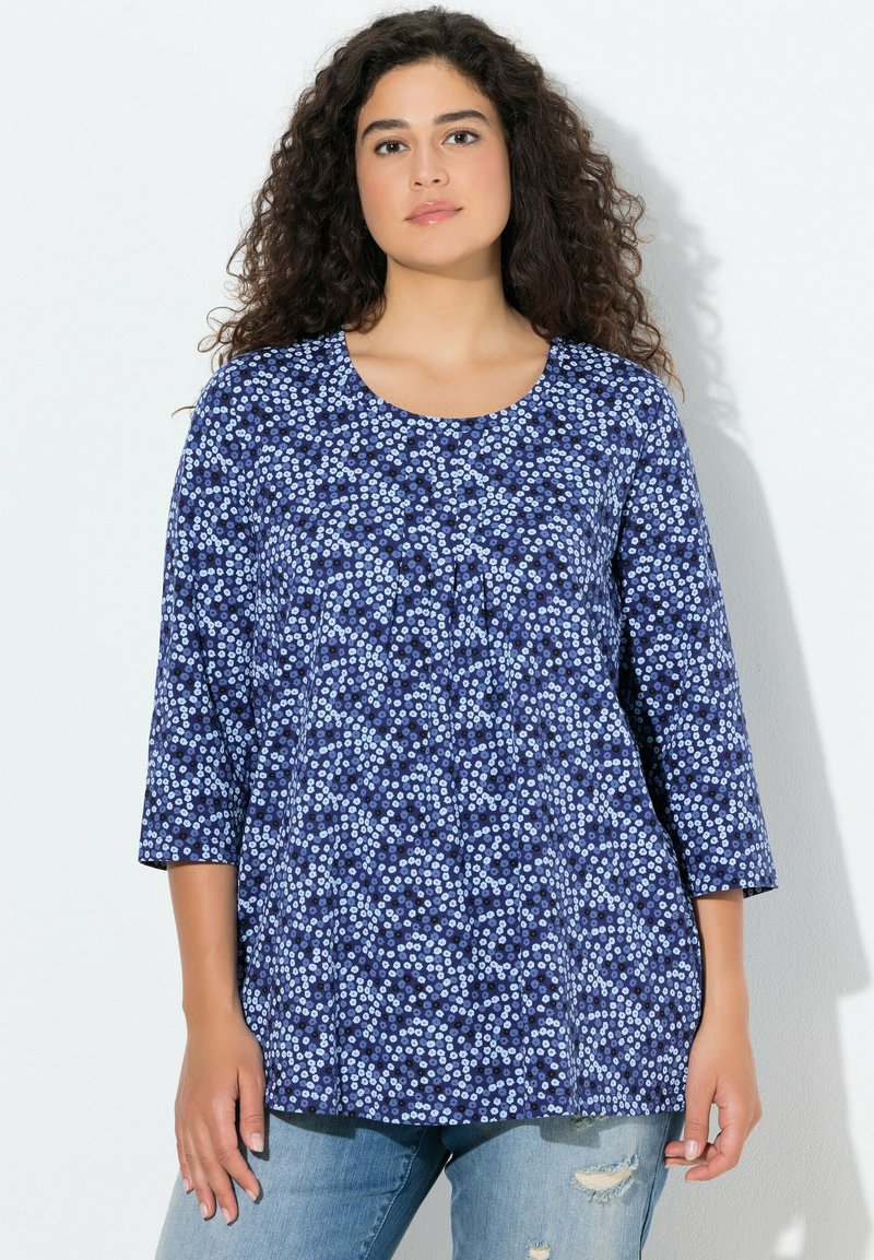 Granatowa bluzka w kwiatowy wzór, z rękawami 3/4, okrągłym dekoltem i plisowanymi detalami z przodu. Noszona na jasnoniebieskich jeansach.