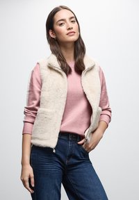 Faux-Fur-Weste in Creme mit Reißverschluss, kombiniert mit einem weichen rosa Pullover und dunkelblauen Jeans. Der Hintergrund ist reinweiß.