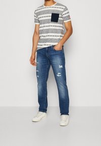 T-shirt a maniche corte a righe bianca e blu navy con taschino sul petto, abbinata a jeans blu strappati e sneaker bianche.