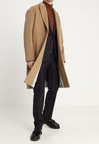 Uomo con cappotto cammello sopra un dolcevita marrone e un completo a scacchi blu, indossa mocassini con frange su uno sfondo bianco semplice.