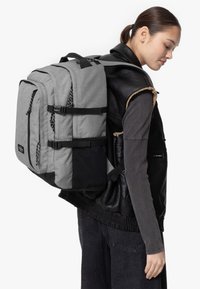 Mochila de tela gris con detalles en negro, múltiples compartimentos, correas laterales y un asa resistente. Llevada en un hombro.