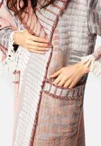 Žena v vzorovanom, frakovanom textilnom kabáte s vreckami, jedna ruka vo vrecku a druhá upravuje predný okraj.