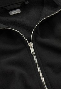 Sort zip-jakke med en glat, struktureret stof. Har en metal lynlås og et mærke med teksten "NEXT." Enkelt, minimalistisk design.