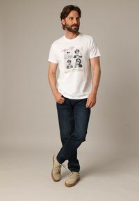 Hombre con barba vistiendo una camiseta blanca con estampado de una calavera, jeans oscuros y zapatillas beige, de pie con una pierna cruzada sobre la otra.