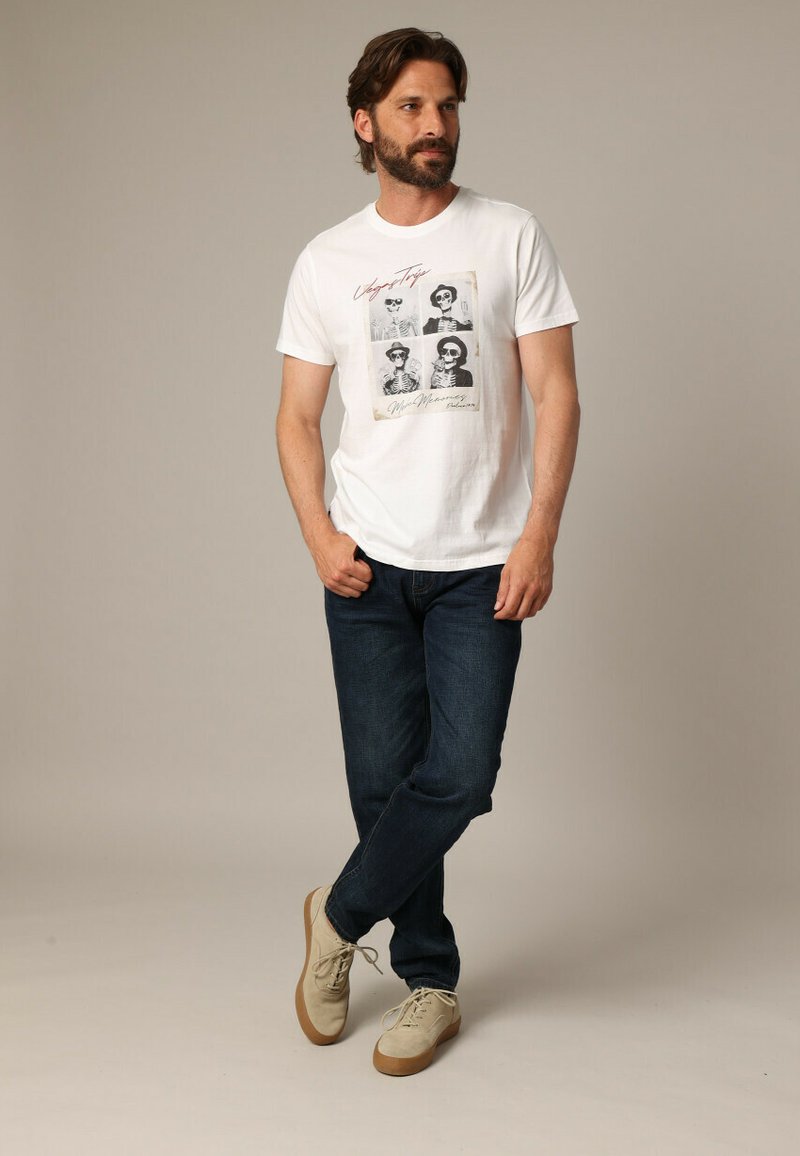 Hombre con barba vistiendo una camiseta blanca con estampado de una calavera, jeans oscuros y zapatillas beige, de pie con una pierna cruzada sobre la otra.