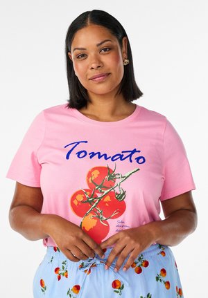 Žena s tamnom kosom srednje dužine nosi ružičastu majicu s crvenim rajčicama i natpisom „Tomato“, uparenu sa svijetloplavim hlačicama s uzorkom rajčica.