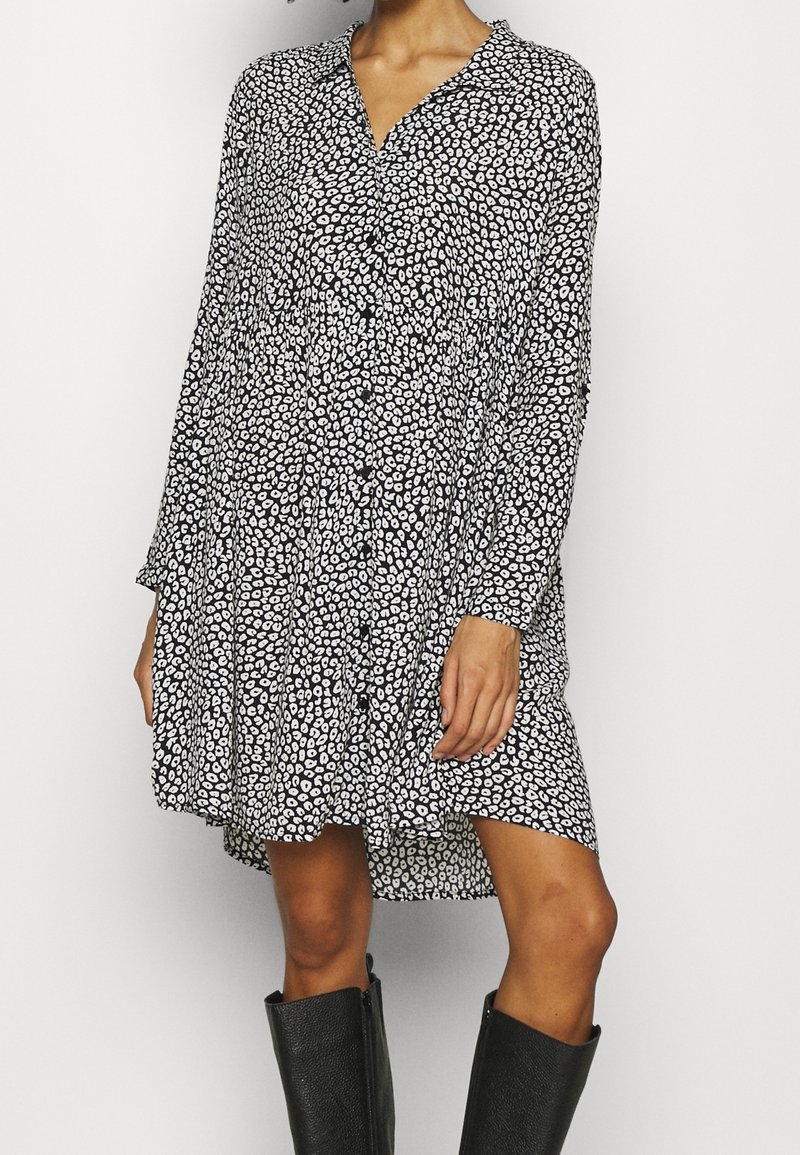 Femme portant une robe noire et blanche à manches longues à motifs tachetés avec un col en V, associée à des bottes noires hautes jusqu'aux genoux, debout devant un fond uni.