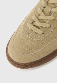 Beżowy zamszowy przód sneakersa z dopasowanymi beżowymi sznurówkami i brązową gumową podeszwą, pokazujący szczegółowe przeszycia i fakturę.