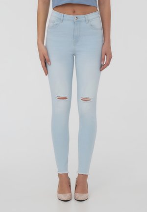 Jeans skinny blu chiaro con dettagli strappati sulle ginocchia, orlo sfilacciato, texture liscia e design tradizionale a cinque tasche. Indossati con tacchi nudi.