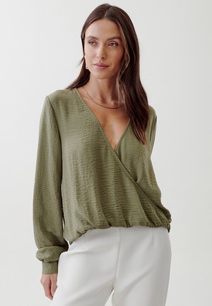Olive green, long-sleeve wrap top with textured fabric, deep V-neckline, and elasticized hem, paired with white trousers.