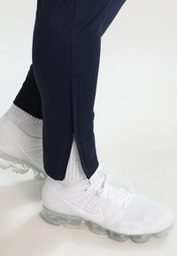 Granatowe spodnie sportowe z zamkiem na kostce, wykonane z elastycznego materiału, zestawione z białymi siateczkowymi sneakersami z przezroczystą podeszwą.