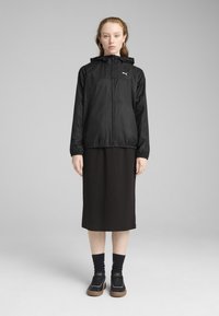 Veste légère noire avec capuche, fermeture éclair avant, poignets élastiques, associée à une robe noire longueur genou et des baskets noires avec semelles marron.