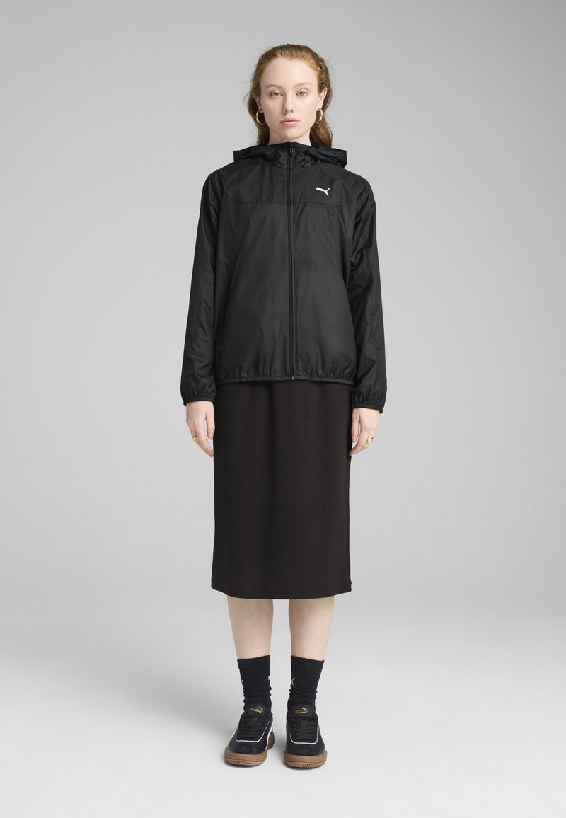 Veste légère noire avec capuche, fermeture éclair avant, poignets élastiques, associée à une robe noire longueur genou et des baskets noires avec semelles marron.