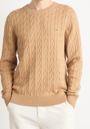 Beżowy sweter w splot warkoczowy z prążkowanymi mankietami i ściągaczem, noszony z jasnymi spodniami, z małym logo Tommy Hilfiger na piersi.