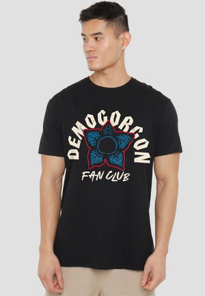Schwarzes Baumwoll-T-Shirt mit einem blauen und roten "Demogorgon Fan Club" Grafik in einer kräftigen, gealterten Schriftart. Kurzärmeliger Schnitt und Rundhalsausschnitt.